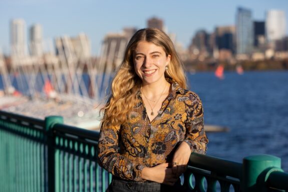 Aviva Intveld, smiling and leaning on a railing overlooking the Charles River with the Boston skyline in the background.