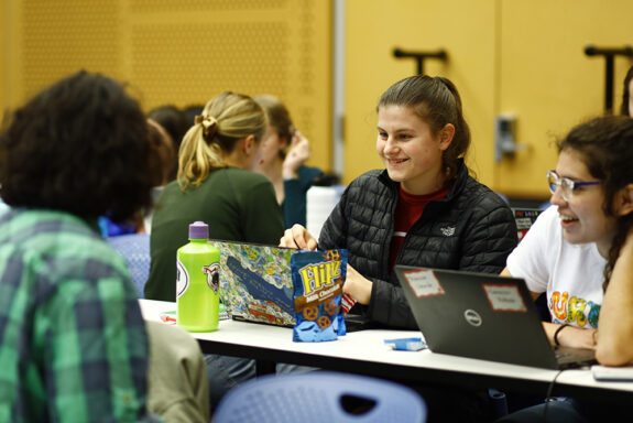 Several MIT students sitting at a narrow table collaborate in a classroom. Two have open laptops and there are snacks on the table.