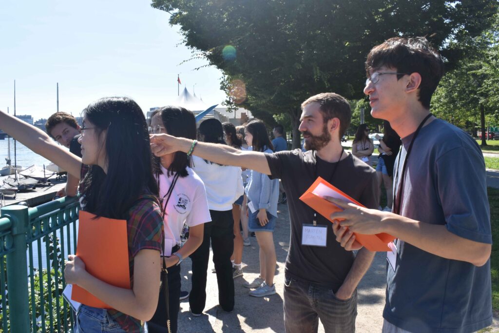 A group of people stand on a bridge, looking in the same direction. Two people in the foreground are pointing at something off caxmera while a third looks on.