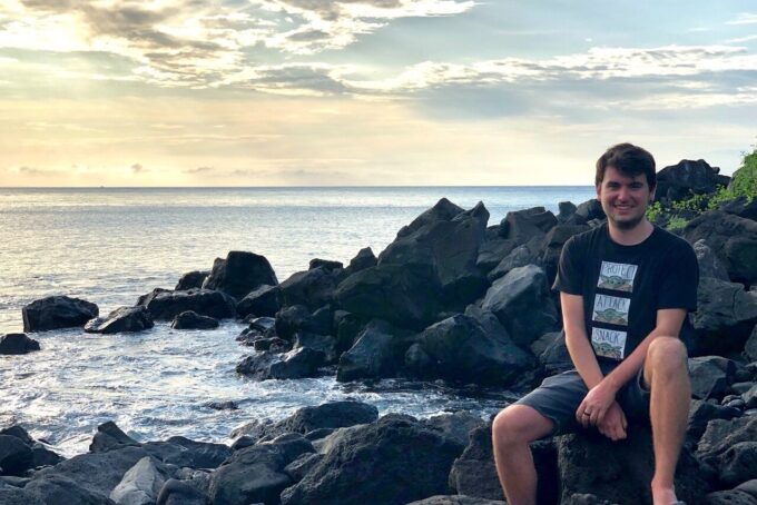 Timur Cinay sits on a rocky outcrop on the sea, smiling at the camera while the sun warms the ocean colors.