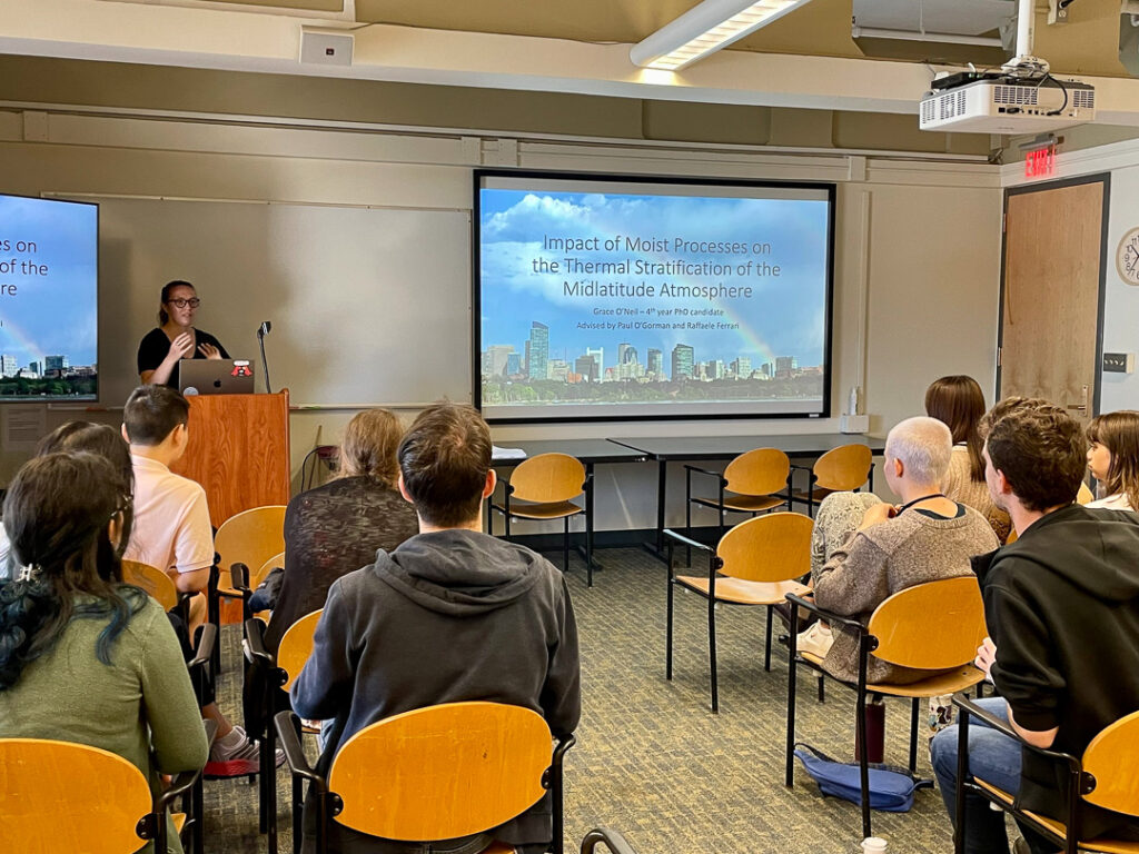 A group of students sit in a small classroom, watching as Grace O'Neil presents a slide labeled "Impact of moist processes on the thermal stratification of the midlatitude atmospshere"