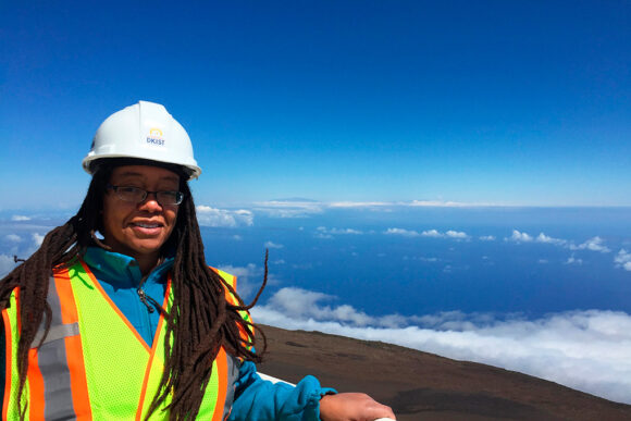 A photo of MIT alum Dara Norman wearing a hard had and a yellow reflective vest with her hand leaning on a railing and a blue sky and landscape behind her.