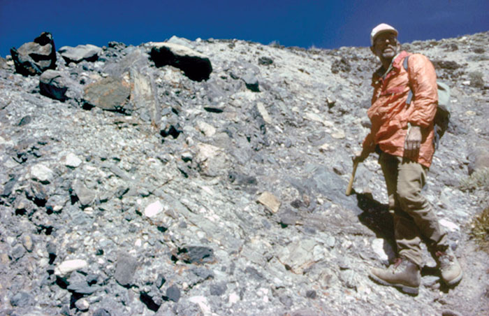 B. Clark Burchfiel stands on a talus field holding a geology hammer.