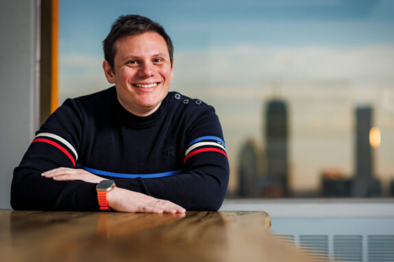 Andrew Babbin sits inside with Boston skyline in background.